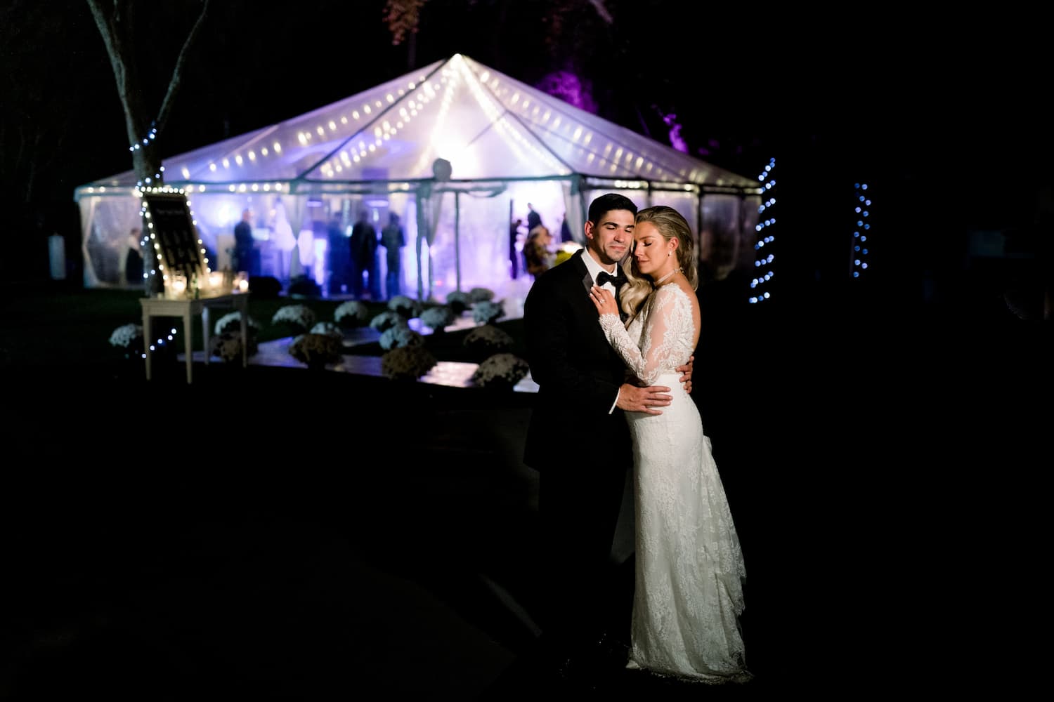 a bride and green infront of a lit up wedding tent